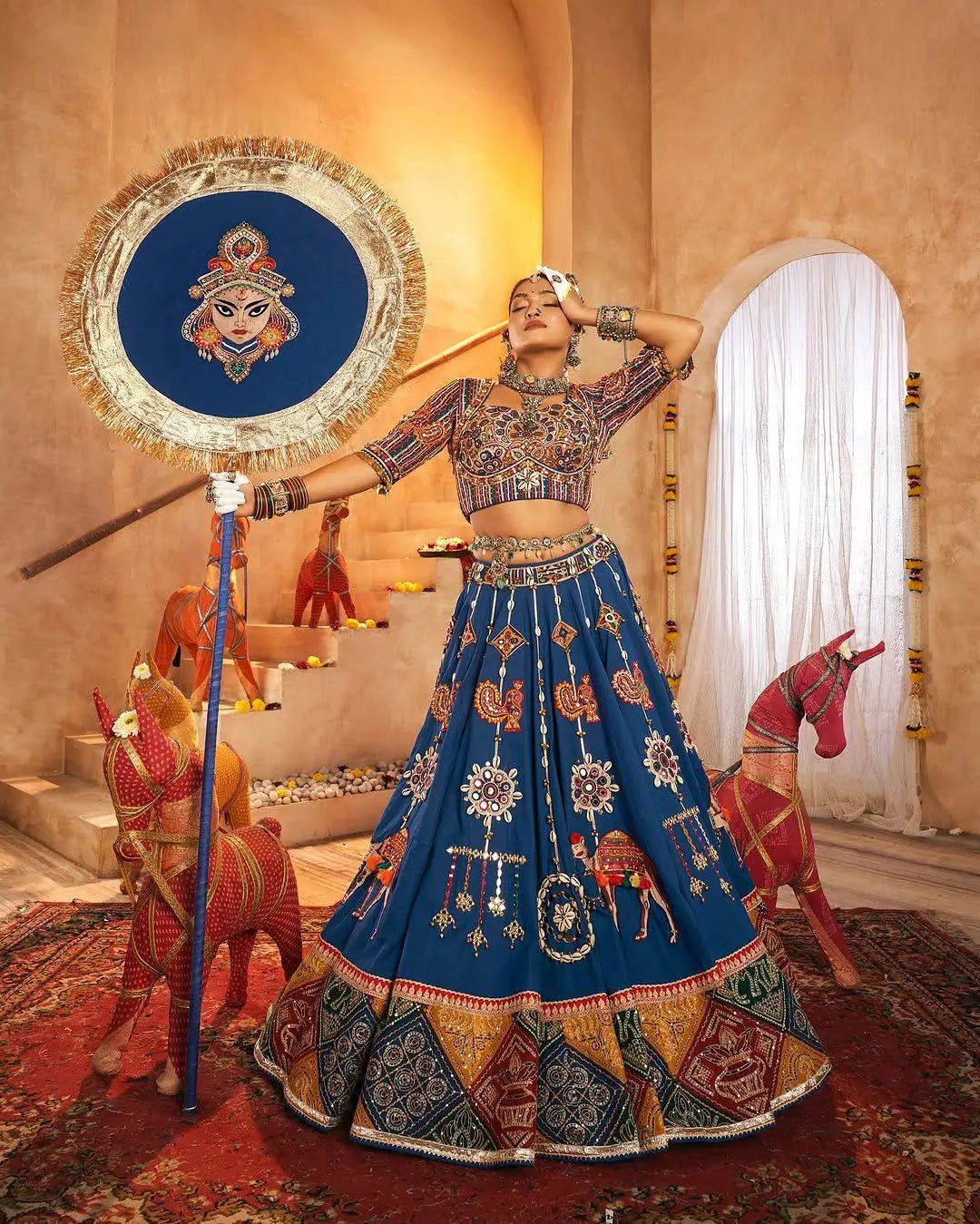 Woman in traditional blue embroidered outfit with decorative elements in a decorated room.